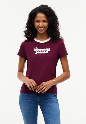 Donna sorridente con capelli ricci che indossa una t-shirt viola Tommy Jeans e jeans blu, in piedi con le mani leggermente giunte davanti.