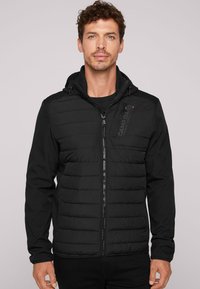 Mustat puhvijakku, jossa on vetoketjukiinnitys edessä, huppu ja teksturoitu yläosa. Sisältää logon rinnassa ja sileää materiaalista hihassa.