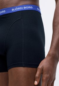 Man wearing black boxer briefs with a blue Björn Borg waistband, shown from the waist to mid-thigh on a white background.
