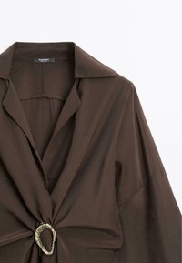 Blouse marron foncé avec un col large, des manches longues et un tissu froncé à travers un anneau argenté irrégulier au centre devant.