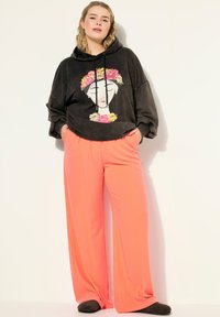 Černá mikina s kapucí a květinovou grafikou na obličeji, kombinovaná s volnými, jasně korálovými širokými kalhotami. Měkká textura, pohodlný střih.