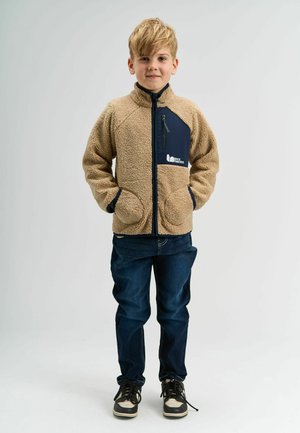 Hnědá fleecová bunda s námořnickými akcenty, propínacím hrudním kapsou, bočními kapsami a žebrovanými manžetami. Nositá přes modré denimové kalhoty a černé boty.