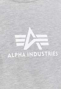 Logo cu un „A” stilizat și trei linii orizontale pe fiecare parte deasupra textului „ALPHA INDUSTRIES” pe o țesătură gri deschis.