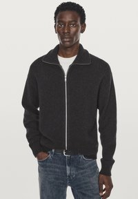 Gray zip-up sweater with a ribbed collar and cuffs, made of textured fabric. Worn over a white shirt, paired with blue jeans.