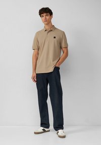 Beige Poloshirt mit einer Drei-Knopf-Leiste und einem kleinen quadratischen Logo. Kombiniert mit dunklen Jeans und weißen Sneakers. Minimalistisches Design.