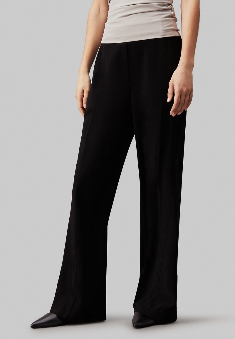 Personne portant un pantalon noir ample à jambes larges, un top beige sans manches et des chaussures noires à bouts pointus, debout sur fond gris.