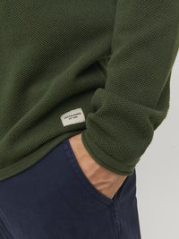 Grønn strikkegenser med teksturert stoff. Har et merke nederst. Kombinert med marineblå bukser som viser lomme detaljer.