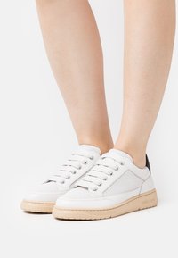 Baskets en cuir perforé blanc avec une semelle en caoutchouc beige clair, dotées de lacets gris et d'un accent noir au talon.