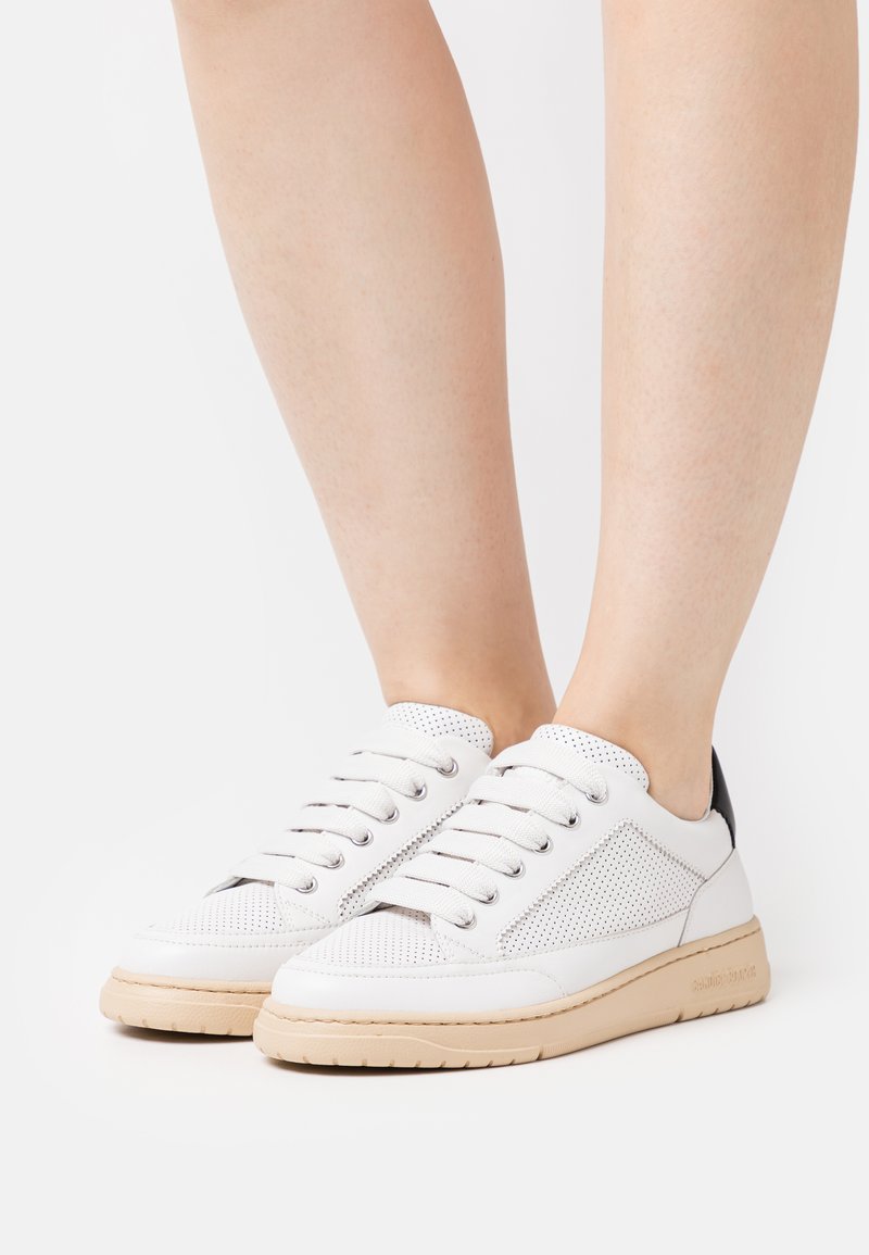 Sneakers bianche in pelle perforata con suola in gomma beige chiaro, dotate di lacci grigi e un accento nero sul tallone.