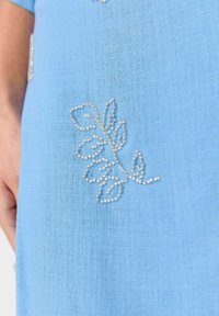 Tissu bleu clair avec un délicat broderie de perles blanches en motif de feuilles, texture lisse, avec une couture visible et une coupe décontractée.