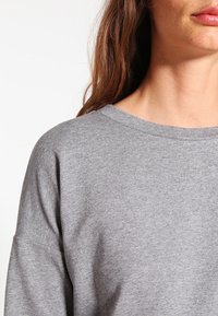 Maglietta grigia in cotone a maniche lunghe con ampio scollo rotondo e vestibilità ampia. Tessuto morbido con cuciture visibili lungo lo scollo e le spalle.