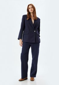 Navyblauwe op maat gemaakte blazer met twee knopen, zijzakken en lange mouwen, gecombineerd met bijpassende wijde broek en bruine schoenen.