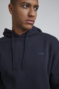 Marineblå hettegenser laget av mykt materiale. Har en frontlomme med kenguru-design, snor til justering, og en subtil "blend"-logo på brystet.