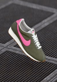 Nike kedas olīvzaļā krāsā ar rozā Swoosh, ar teksturētu virsmu, baltām auklām un brūnu gumijas zoli.