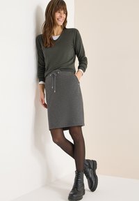 Jupe grise à cordon avec un tissu texturé, associée à un haut à manches longues vert foncé. Des bottes noires à lacets complètent la tenue.