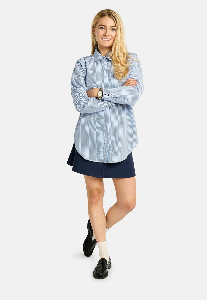 Mėlynos ir baltos spalvos dryžuota ilgomis rankovėmis marškiniai su sagomis, derinami su tamsiai mėlyna suknele, juodais laisvalaikio batais ir baltais kojinaitėmis. Blizgus sidabrinis laikrodžio detales.