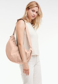 Borsa a spalla beige realizzata in morbido tessuto, con una forma arrotondata, tracolla regolabile e dettagli hardware discreti. Indossata da una modella.