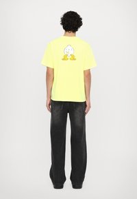 T-shirt giallo neon con una grafica di un pulcino in stile cartoon sulla schiena. Indossato con jeans scuri a gamba larga. Il tessuto appare morbido e informale.