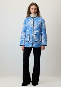 Plavi jastučić s cvjetnim uzorkom u bijeloj boji, crnim obrubom i velikim prednjim džepovima. Nošen preko visokog ovratnika i crnih širokih hlača.