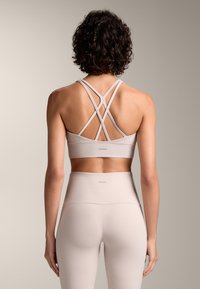 Ljus beige sport-bh med korsade axelband och matchande högmidjade leggings. Slät textur, minimalistisk design, märkeslogotyp på bandet.