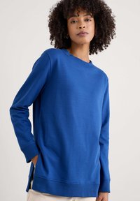 Kék pulóver kerek nyakkivágással, hosszú ujjakkal és oldalsó részekkel. Simább textúrával és bordás mandzsettákkal, valamint az alján.