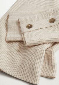 Beige ribbattuina neulekangasta, jossa on kaksi nappia taitetussa hihassa, esittäen teksturoitua pintaa ja siistejä ompelutietoja.