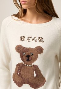 Kobieta w białym swetrze z brązowym wzorem niedźwiadka oraz napisem "BEAR" ułożonym z dekoracyjnych kamieni na piersi.