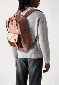 Sac à dos avec un extérieur rose, une poche avant beige et un fond marron. Il dispose de bretelles réglables, de matériel doré et d'un écusson logo rectangulaire.