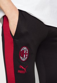 Schwarze Hose mit roten Seitenstreifen, ausgestattet mit gesticktem AC Milan-Logo und Puma-Branding. Glatte Textur, seitliche Tasche sichtbar.