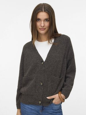 Brauner Wollcardigan mit V-Ausschnitt, Knopfverschluss, zwei Taschen vorn, gerippten Bündchen und Saum. Getragen über einem weißen Oberteil und Jeans.