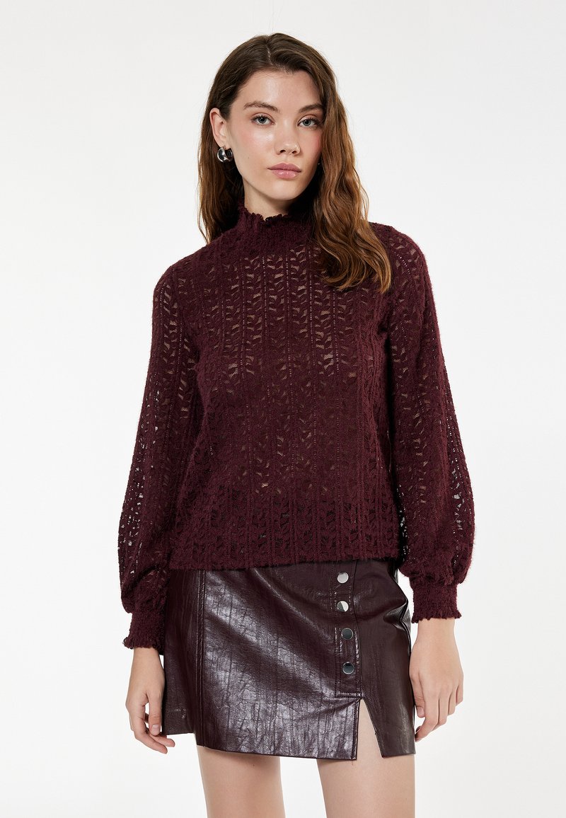 Maglione lavorato a maglia bordeaux con collo alto e maniche a sbuffo, caratterizzato da un motivo a rete; abbinato a una minigonna in pelle coordinata.