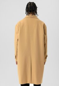 Lange beige jas gemaakt van gestructureerde stof met een klassieke kraag, met knopen sluitbare manchetten en een centrale achternaad, ontworpen voor een losse pasvorm.