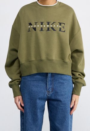 Person trägt ein olivgrünes, kurzes Sweatshirt mit dem Logo "NIKE SPORTSWEAR" und blaue Jeans mit hoher Taille, steht vor einem einfarbigen Hintergrund.