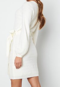 Femme portant une robe en maille blanche avec des manches longues bouffantes et une ceinture nouée, vue de profil sur un fond blanc uni.