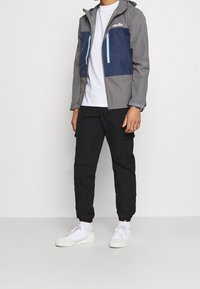 Jasnoszara wodoodporna kurtka z granatowymi przednimi panelami, regulowanym kapturem, kieszeniami na zamek, noszona na białej koszuli i czarnych spodniach. Białe buty.