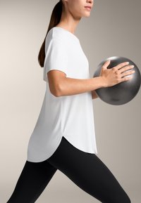 Bílý krátký rukáv top s zaobleným lemem, skombinovaný s černými legínami. Postava drží tmavě šedou cvičební kouli, což demonstruje aktivní postoj.