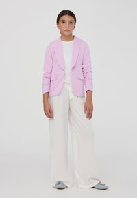 Blazer rosa chiaro con colletto a scialle e due tasche frontali, abbinato a pantaloni bianchi a gamba larga e scarpe blu slip-on.