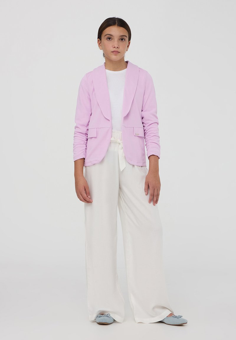 Blazer rosa chiaro con colletto a scialle e due tasche frontali, abbinato a pantaloni bianchi a gamba larga e scarpe blu slip-on.