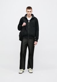 Μαύρο τζάκετ με φερμουάρ και ριμπ μανσέτες, συνδυασμένο με μαύρο παντελόνι με φαρδύ πόδι. Το μοντέλο φοράει λευκά αθλητικά και κρατάει μαύρη τσάντα. Ουδέτερο φόντο.