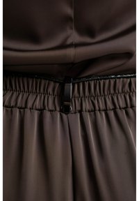 Brązowa, gładka tkanina z marszczoną talią oraz połyskującą czarną klamrą. Szczegóły obejmują cienki czarny akcent wzdłuż talii.