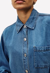 Blauwe denim overhemd met een knoopsluiting kraag, zilverkleurige hardware en een borstzak. Kenmerkt zichtbare stiksels en een gladde textuur.