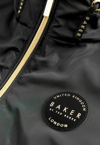 Giacca nera con zip dorata e un patch rotondo nero con scritte dorate: "BAKER by TED BAKER" e "REGNO UNITO," "LONDRA."