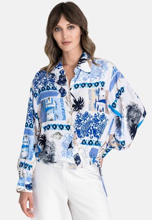 Vrouw die een losse blauw-wit patroon blouse met knopen en lange mouwen draagt, gecombineerd met een witte broek, tegen een effen achtergrond.