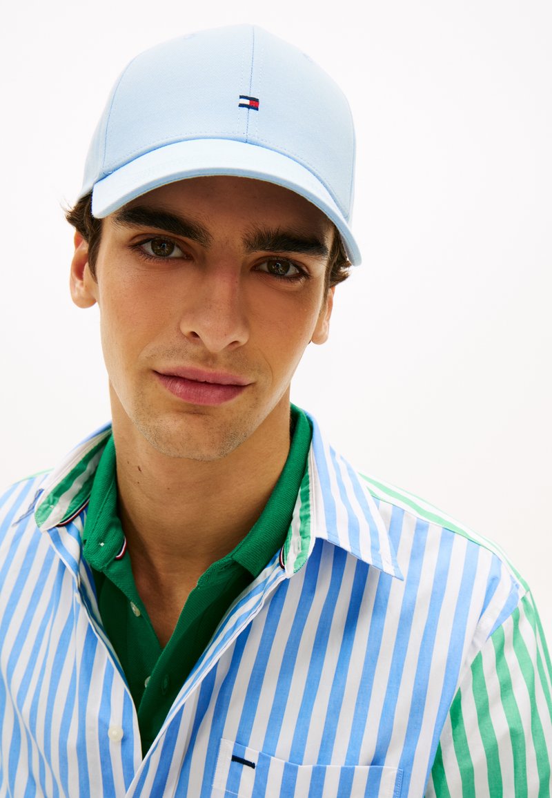 Ανοιχτό μπλε καπέλο μπέιζμπολ με κεντημένο λογότυπο, φορεμένο με πράσινο πόλο πουκάμισο και ριγέ πουκάμισο με κουμπιά, εμφανείς λεπτομέρειες γιακά και υφές.