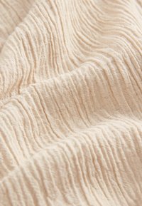 Skrynkligt tyg i en mjuk beige nyans, som visar på en fin, upphöjd struktur med horisontella linjer genom hela. Lätt och luftigt material.
