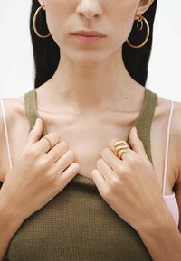 Pendientes de aro de oro, una camiseta sin mangas de punto verde oliva texturizada y dos anillos: uno con banda lisa y otro con un diseño retorcido y orgánico.