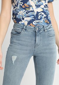 Jeans de mezclilla azul claro con un corte ajustado, que presentan una zona de rodilla desgastada y un estilo clásico de cinco bolsillos. Cierre de botón en la cintura.