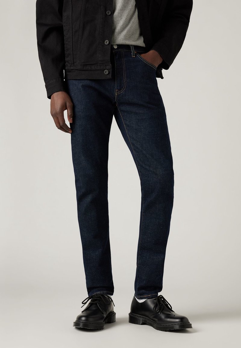 Pantalones vaqueros de mezclilla azul oscuro con un corte recto, detalles de costura y diseño de cinco bolsillos, combinados con una chaqueta negra y zapatos negros.