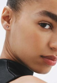 Pendiente de estrella plateada en forma de estrella en la oreja izquierda; la piel suave con un ligero brillo contrasta con una prenda oscura y texturizada.