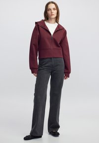 Sudadera corta de color burdeos con capucha y cremallera frontal, combinada con jeans de pierna ancha de alta cintura en gris oscuro. La textura es suave; los materiales son blandos.